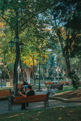 A serene park setting with several wooden benches placed along paved pathways. Tall trees with lush green foliage border the paths, allowing dappled sunlight to filter through. People are seated on the benches, enjoying the peaceful surroundings. A couple is engaged in a conversation on the front bench, adding to the tranquil atmosphere. Vintage-style street lamps line the walkway, enhancing the overall charm of the scene.