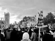 A large group of people is gathered in an urban area, with many holding signs and banners with messages related to Palestine. The background features historic architecture, likely a well-known landmark, under a cloudy sky. Some of the attendees are wearing headscarves and the atmosphere appears to be a public demonstration or protest.