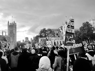 A large group of people is gathered in an urban area, with many holding signs and banners with messages related to Palestine. The background features historic architecture, likely a well-known landmark, under a cloudy sky. Some of the attendees are wearing headscarves and the atmosphere appears to be a public demonstration or protest.