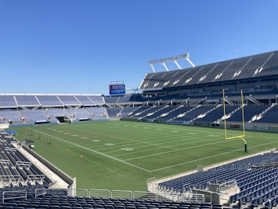A large, empty sports stadium with blue seats surrounding a well-maintained green grass field. Bright sunlight creates clear shadows, and there are two goal posts on opposite ends of the field. A big electronic scoreboard displays some graphics, and the sky is clear and blue.