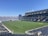 A large, empty sports stadium with blue seats surrounding a well-maintained green grass field. Bright sunlight creates clear shadows, and there are two goal posts on opposite ends of the field. A big electronic scoreboard displays some graphics, and the sky is clear and blue.