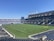 A large, empty sports stadium with blue seats surrounding a well-maintained green grass field. Bright sunlight creates clear shadows, and there are two goal posts on opposite ends of the field. A big electronic scoreboard displays some graphics, and the sky is clear and blue.