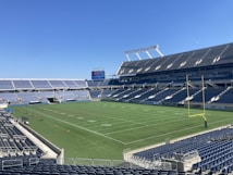 A large, empty sports stadium with blue seats surrounding a well-maintained green grass field. Bright sunlight creates clear shadows, and there are two goal posts on opposite ends of the field. A big electronic scoreboard displays some graphics, and the sky is clear and blue.