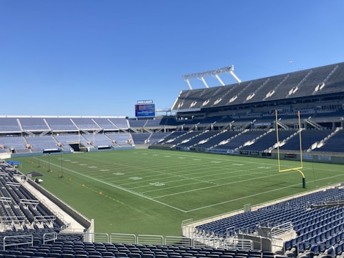 A large, empty sports stadium with blue seats surrounding a well-maintained green grass field. Bright sunlight creates clear shadows, and there are two goal posts on opposite ends of the field. A big electronic scoreboard displays some graphics, and the sky is clear and blue.