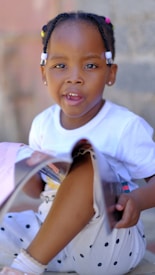 A young child with braided hair adorned with colorful beads sits on the ground. The child wears a white shirt and polka-dotted pants and is holding an open magazine.