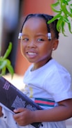 A child with braided hair adorned with white beads is smiling warmly while holding a book. The child is wearing a white shirt with colorful stripes and is surrounded by green leaves.