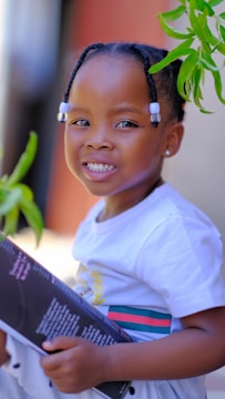 A child with braided hair adorned with white beads is smiling warmly while holding a book. The child is wearing a white shirt with colorful stripes and is surrounded by green leaves.