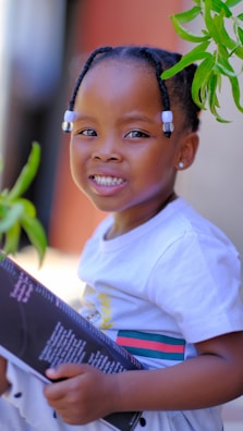 A child with braided hair adorned with white beads is smiling warmly while holding a book. The child is wearing a white shirt with colorful stripes and is surrounded by green leaves.