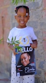 A young child wearing a white t-shirt with a bee design and patterned pants smiles while holding a lifestyle magazine featuring a woman on the cover. The background consists of a stone wall and there are green leaves hanging in the foreground.