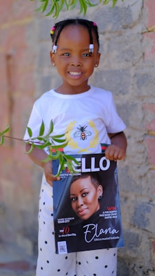 A young child wearing a white t-shirt with a bee design and patterned pants smiles while holding a lifestyle magazine featuring a woman on the cover. The background consists of a stone wall and there are green leaves hanging in the foreground.