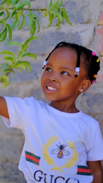 A young child with beaded hair reaches towards green leaves. The child is wearing a white shirt featuring a bee emblem.