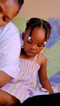 A young child with braided hair adorned with beads engages in an activity while sitting close to an adult. The child wears a colorful, striped dress and is focused intently on something out of view. The background shows soft, blurred colors, adding a gentle ambiance to the scene.