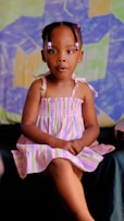 A young child wearing a striped dress sits with her hands folded on her lap against a colorful, geometric-patterned background. Her hair is styled with beads, and she appears to be in a relaxed pose.