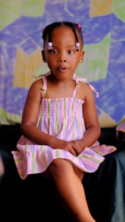 A young child wearing a striped dress sits with her hands folded on her lap against a colorful, geometric-patterned background. Her hair is styled with beads, and she appears to be in a relaxed pose.