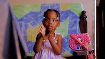 A young child in a colorful striped dress is sitting and clapping. The background features a colorful abstract painting with geometric shapes. Beside the child is a small bag with a doll illustration on it, decorated with butterflies.