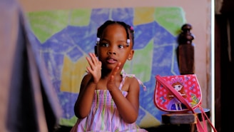 A young child in a colorful striped dress is sitting and clapping. The background features a colorful abstract painting with geometric shapes. Beside the child is a small bag with a doll illustration on it, decorated with butterflies.