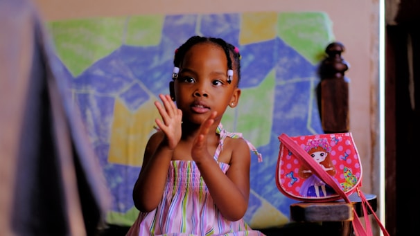 A young child in a colorful striped dress is sitting and clapping. The background features a colorful abstract painting with geometric shapes. Beside the child is a small bag with a doll illustration on it, decorated with butterflies.