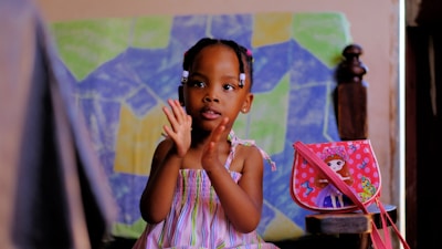 A young child in a colorful striped dress is sitting and clapping. The background features a colorful abstract painting with geometric shapes. Beside the child is a small bag with a doll illustration on it, decorated with butterflies.