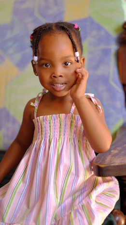 A young child with braided hair adorned with colorful beads is sitting on a wooden chair. She is wearing a pastel-striped dress and is posing with her hand near her face. The background features a geometric pattern in soft colors.