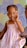 A young child with braided hair adorned with colorful beads is sitting on a wooden chair. She is wearing a pastel-striped dress and is posing with her hand near her face. The background features a geometric pattern in soft colors.