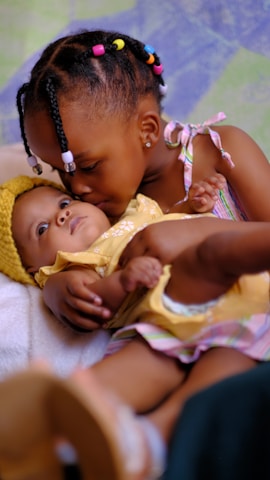A young child gently kisses a baby who is wearing a yellow outfit and hat while being cradled in their arms. The setting is warm and intimate, with a soft background featuring pastel shades of blue and green. The child's hair is styled with colorful beads.