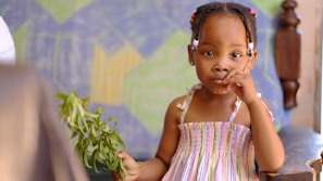 A young child with braided hair, adorned with pink and white beads, wearing a colorful striped dress, holds a branch with green leaves while sitting on a wooden chair. The background includes a geometric patterned wall with shades of blue, green, and yellow.