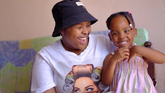 A person wearing a white t-shirt with a graphic print and a black bucket hat is sitting next to a young child in a colorful dress. Both are smiling and appear to be happy, conveying a sense of joy and togetherness. The background includes a multicolored blanket or fabric.