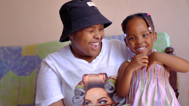 A person wearing a white t-shirt with a graphic print and a black bucket hat is sitting next to a young child in a colorful dress. Both are smiling and appear to be happy, conveying a sense of joy and togetherness. The background includes a multicolored blanket or fabric.