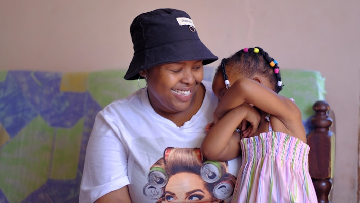 A woman wearing a black bucket hat and a white t-shirt sits on a patterned sofa, smiling and holding a young girl. The child, with colorful beads in her hair, leans affectionately against the woman. The atmosphere appears warm and familial.