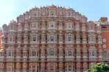 A grand architectural structure featuring numerous small windows with intricate latticework and a symmetrical façade. The building exhibits traditional design elements typical of historic Indian architecture, with a warm terracotta color contrasting against the clear blue sky.
