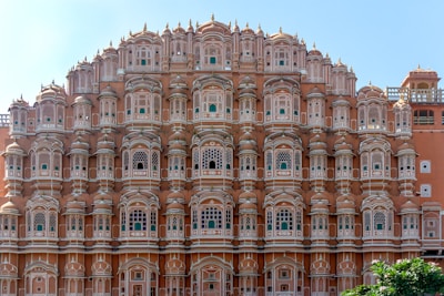 A grand architectural structure featuring numerous small windows with intricate latticework and a symmetrical façade. The building exhibits traditional design elements typical of historic Indian architecture, with a warm terracotta color contrasting against the clear blue sky.