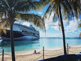 A massive cruise ship is docked near a tropical beach, where palm trees frame the scene. The water is a clear, inviting turquoise, and a few people are swimming. A person sits on the sandy beach, enjoying the sunny day.