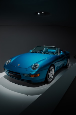 A sleek, blue sports car is positioned against a dark backdrop, illuminated dramatically from above. The car features a convertible design with smooth, aerodynamic lines. The headlights are round, and the brand logo is visible on the front. It is displayed on a white surface that reflects minimal light, emphasizing its vibrant color.