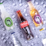 Various soda bottles are arranged on a bed of ice cubes. The central focus is a Coca-Cola bottle, surrounded by Thomas Henry beverages and a bottle of Adelholzener. Each bottle is covered with droplets, giving a chilled appearance.