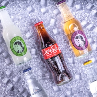 Various soda bottles are arranged on a bed of ice cubes. The central focus is a Coca-Cola bottle, surrounded by Thomas Henry beverages and a bottle of Adelholzener. Each bottle is covered with droplets, giving a chilled appearance.