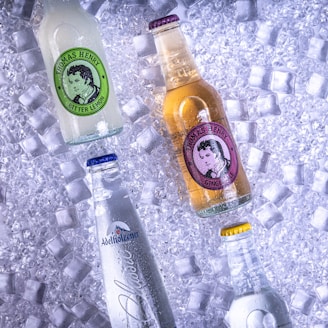 Several bottles of beverages are placed on a bed of ice cubes. The bottles have different labels and colors, with distinct brand names visible on each. One is a brownish liquid labeled 'Thomas Henry Ginger Ale', another is a clear liquid labeled 'Adelholzener', while another has a green label.