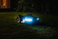 A robotic lawn mower with LED lights is operating on a grassy lawn during the night. The device is sleek and modern, emitting a blue glow that contrasts with the dark surroundings. In the background, soft ambient light appears from a window of a house.