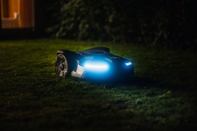 A robotic lawn mower with LED lights is operating on a grassy lawn during the night. The device is sleek and modern, emitting a blue glow that contrasts with the dark surroundings. In the background, soft ambient light appears from a window of a house.