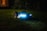 A robotic lawn mower with LED lights is operating on a grassy lawn during the night. The device is sleek and modern, emitting a blue glow that contrasts with the dark surroundings. In the background, soft ambient light appears from a window of a house.