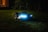 A robotic lawn mower with LED lights is operating on a grassy lawn during the night. The device is sleek and modern, emitting a blue glow that contrasts with the dark surroundings. In the background, soft ambient light appears from a window of a house.
