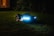 A robotic lawn mower with LED lights is operating on a grassy lawn during the night. The device is sleek and modern, emitting a blue glow that contrasts with the dark surroundings. In the background, soft ambient light appears from a window of a house.