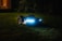 A robotic lawn mower with LED lights is operating on a grassy lawn during the night. The device is sleek and modern, emitting a blue glow that contrasts with the dark surroundings. In the background, soft ambient light appears from a window of a house.