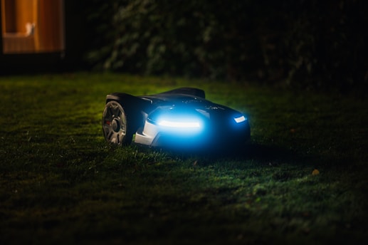 A robotic lawn mower with LED lights is operating on a grassy lawn during the night. The device is sleek and modern, emitting a blue glow that contrasts with the dark surroundings. In the background, soft ambient light appears from a window of a house.