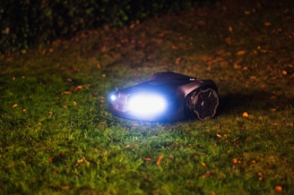 A lawn robot with bright headlights is illuminated in the night, navigating over a grassy area scattered with a few fallen leaves. The background is dimly lit and appears to be a hedge or bush.