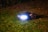A lawn robot with bright headlights is illuminated in the night, navigating over a grassy area scattered with a few fallen leaves. The background is dimly lit and appears to be a hedge or bush.
