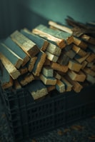 A pile of thin, rectangular wooden planks is stacked inside a black plastic crate. The planks vary in size and appear to be roughly cut. The lighting gives the wood a slightly warm tone, while the background is softly out of focus.