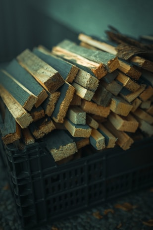 A pile of thin, rectangular wooden planks is stacked inside a black plastic crate. The planks vary in size and appear to be roughly cut. The lighting gives the wood a slightly warm tone, while the background is softly out of focus.