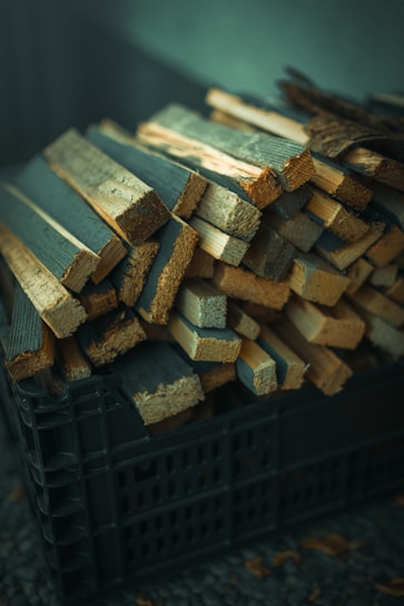 A pile of thin, rectangular wooden planks is stacked inside a black plastic crate. The planks vary in size and appear to be roughly cut. The lighting gives the wood a slightly warm tone, while the background is softly out of focus.
