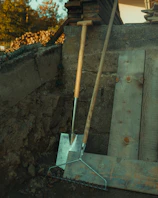 A set of durable landscaping rakes and shovels leaning against a wooden fence.