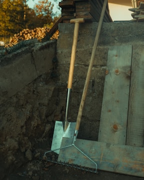 Two wooden-handled gardening tools, a shovel and a rake, are leaning against a concrete wall. Beside them, several wooden planks are stacked. The scene is outdoors, with grass growing near the edges and some trees visible in the blurred background.